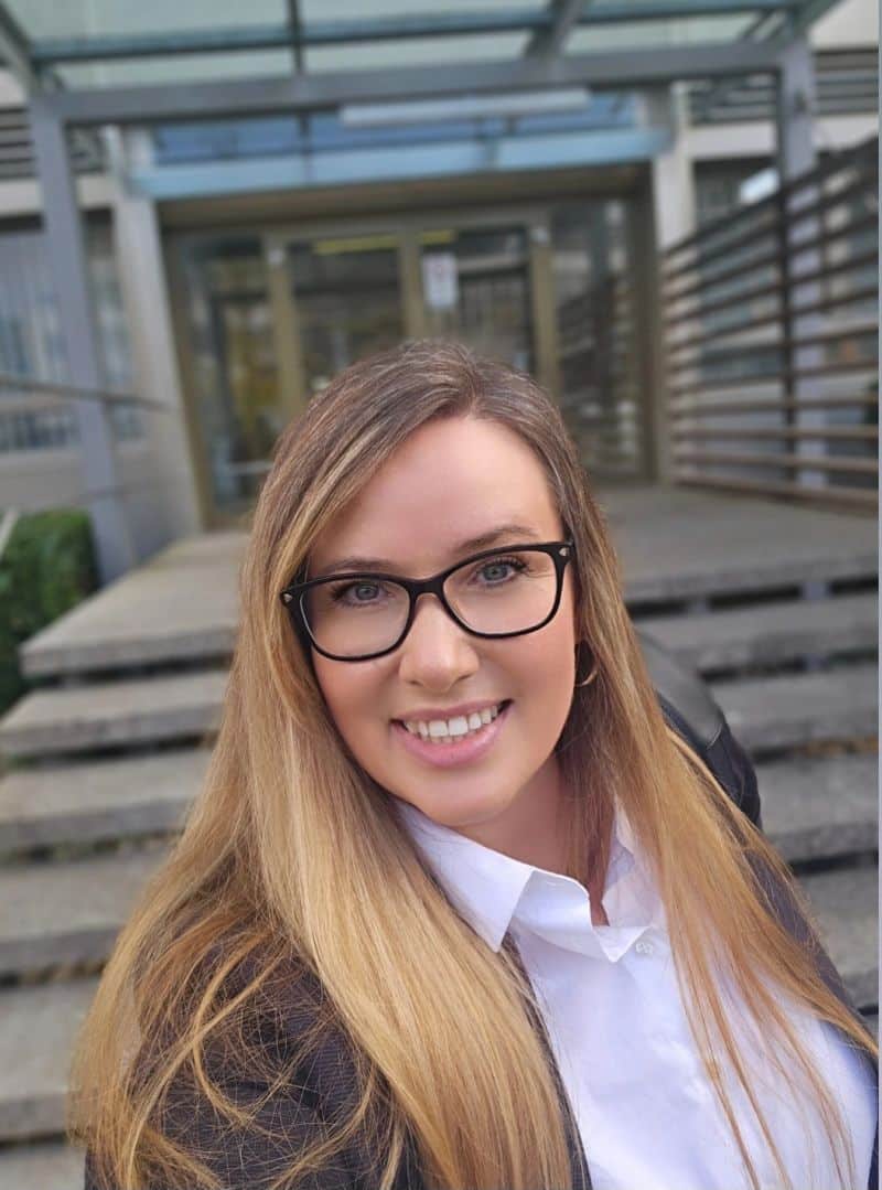 Frau mit langen Haaren und Brille steht vor dem Eingang eines Gebäudes der IHK.